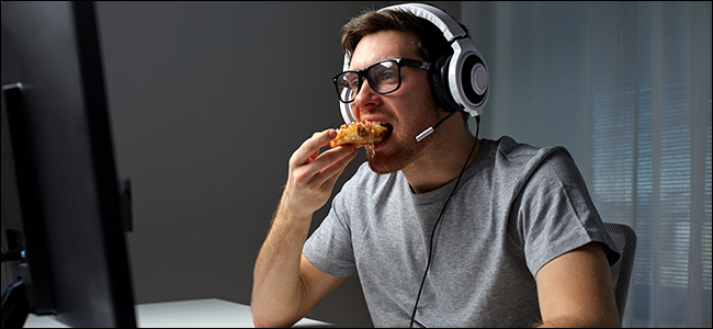 Seorang pria duduk di depan komputer, mengenakan headset, dan makan pizza.