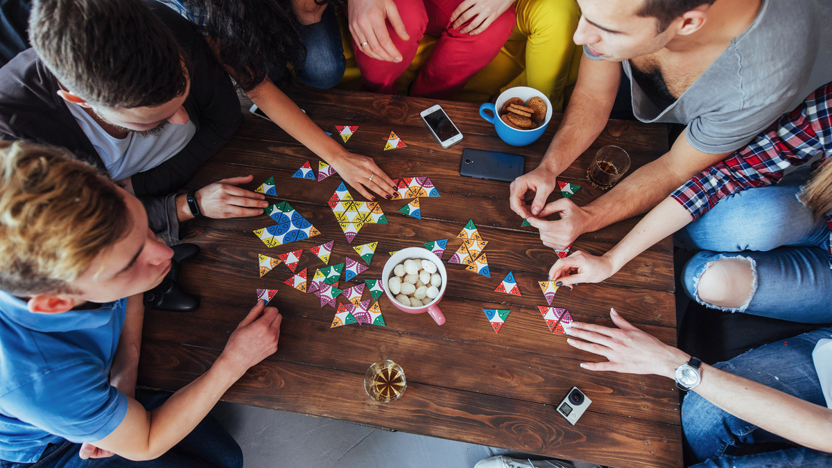 Góc nhìn từ trên xuống của bức ảnh sáng tạo về những người bạn ngồi trên bàn gỗ chơi trò chơi board với đồ ăn nhẹ và đồ uống