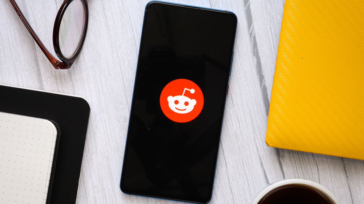 Hình ảnh lưu trữ logo ứng dụng Reddit trên điện thoại Android ngồi trên bàn với cà phê, kính và các phụ kiện khác