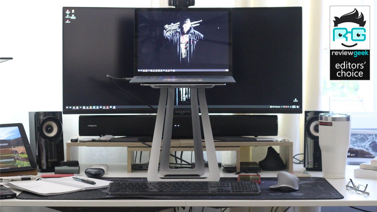 Dudukan laptop seluler VariDesk dengan 3 laptop permukaan di atasnya, duduk di depan layar besar