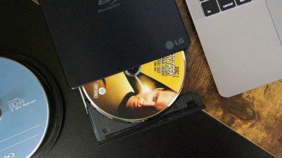 Một trục xoay của Đĩa Blu-ray trống, ổ ghi đĩa quang bên ngoài và MacBook Pro nằm trên bàn làm việc.