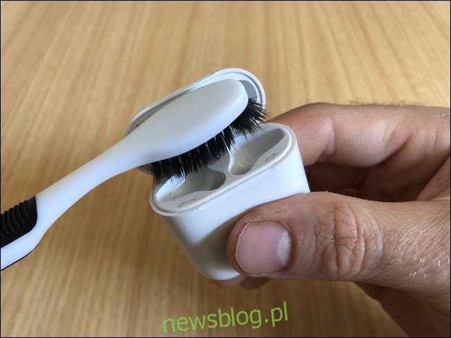 Một tay cầm một cái hộp Apple AirPods và làm sạch chúng bằng bàn chải đánh răng.